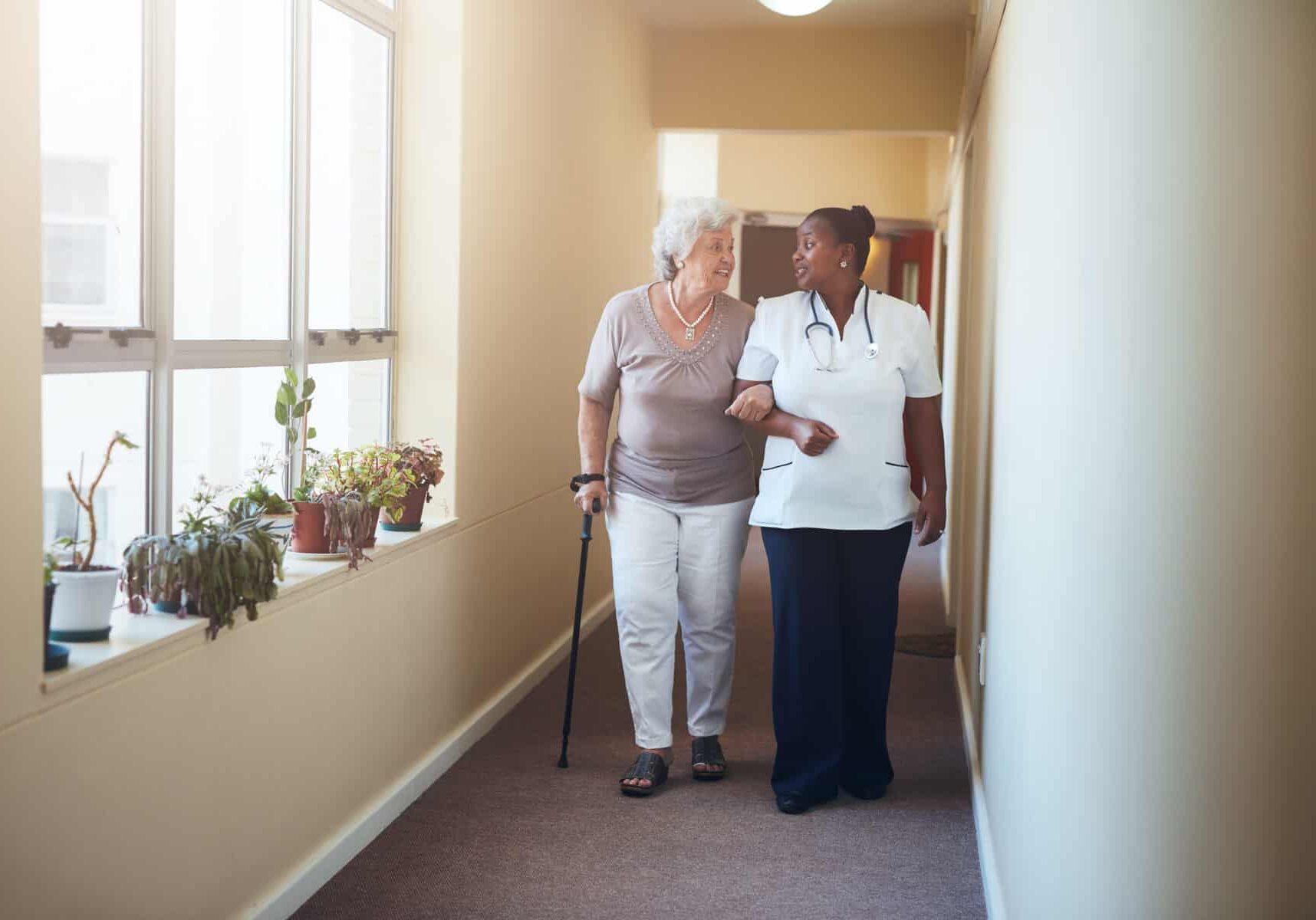 Full length portrait of senior woman walking with her nurse at nursing home. Healthcare work helping female patient.